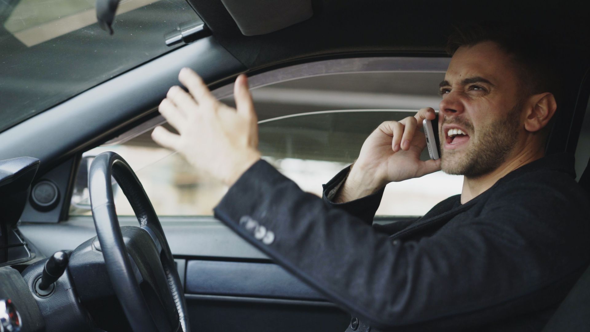 Stressed man gesturing while on phone in car, expressing frustration.