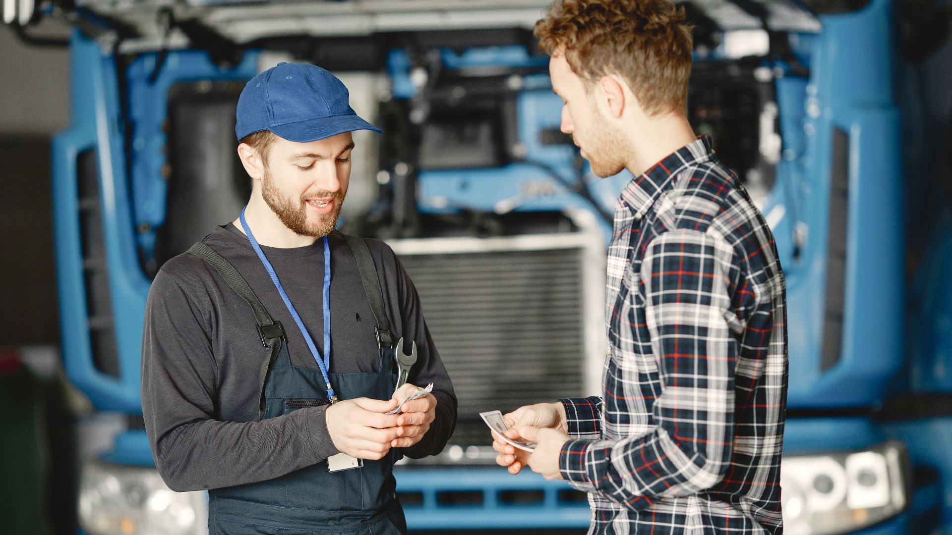 Auto mechanic in blue uniform receives payment from customer in a garage setting with a truck in the background.