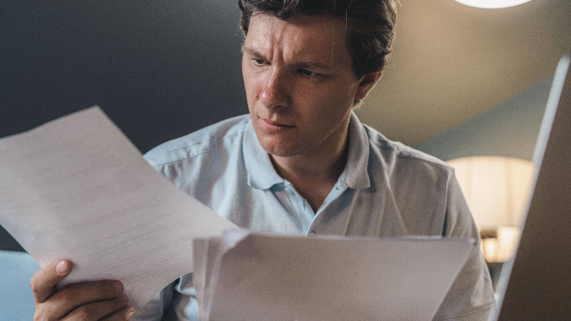 A man intently examines papers, seated indoors under warm lighting, focusing on his work.