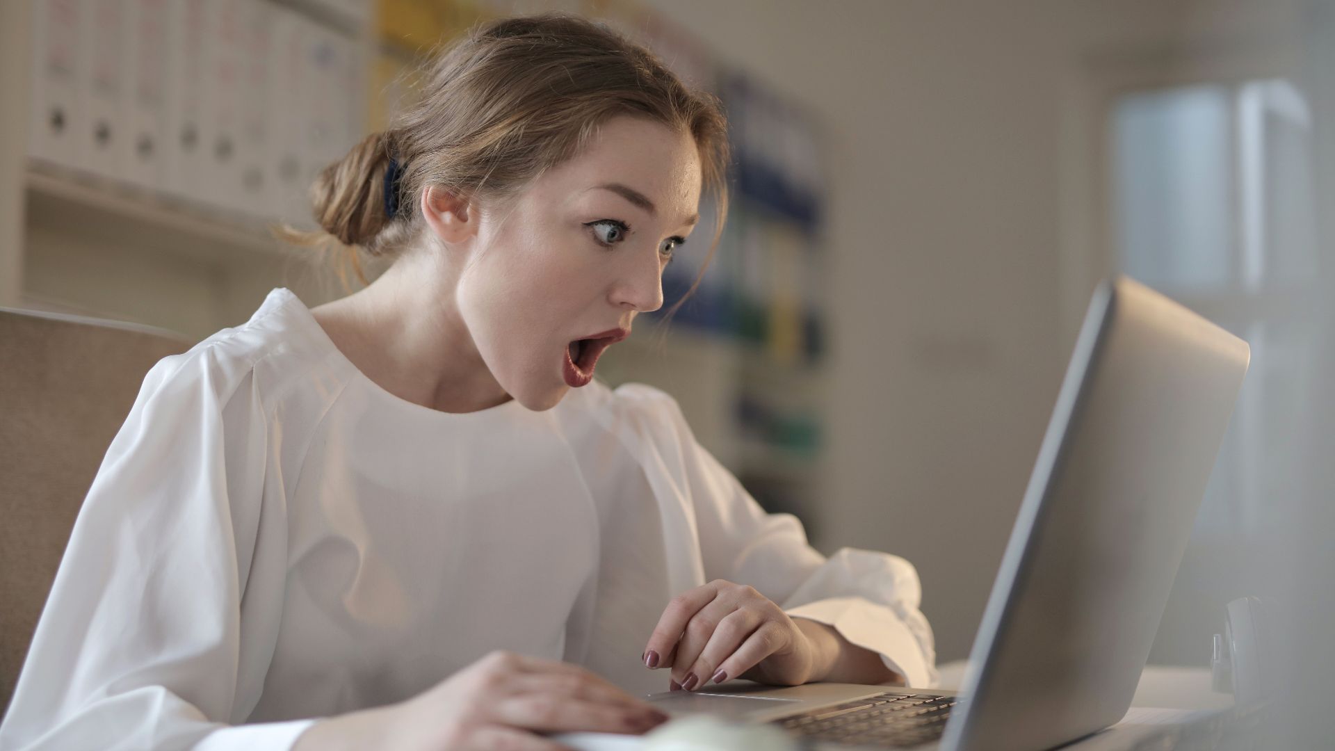 Surprised woman sitting at desk with laptop indoors, expressing amazement.