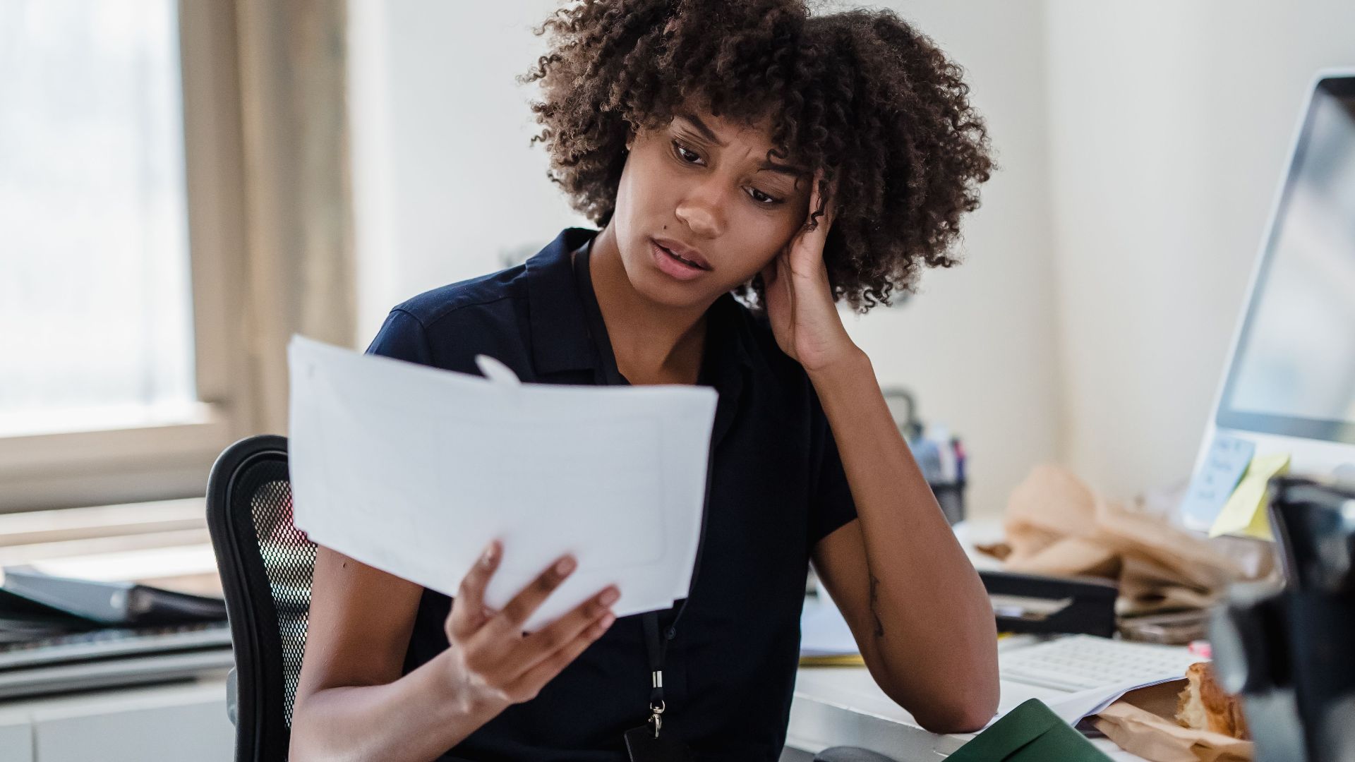 A professional woman with curly hair reviews documents at her office desk, deep in thought.