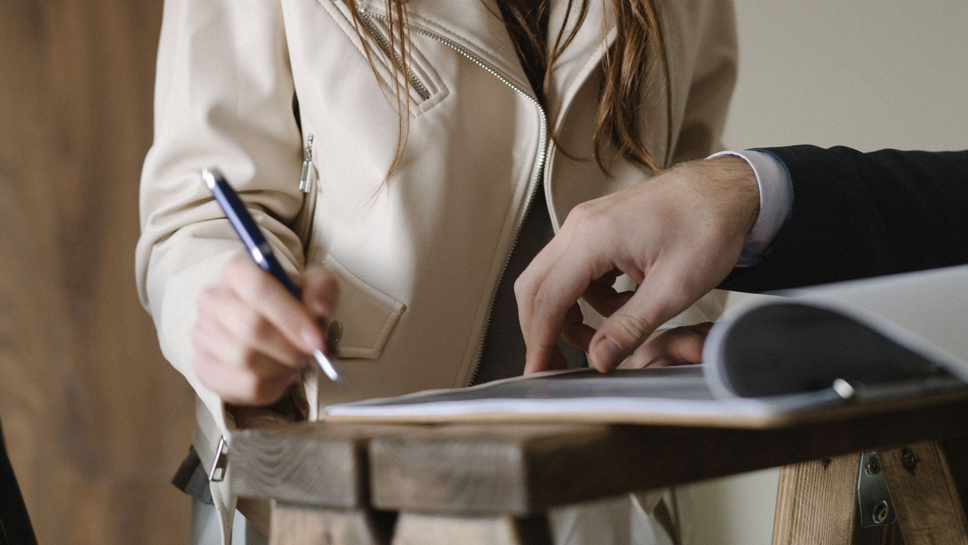 A young woman signing a business contract at an indoor table, holding a pen with focus and intent.