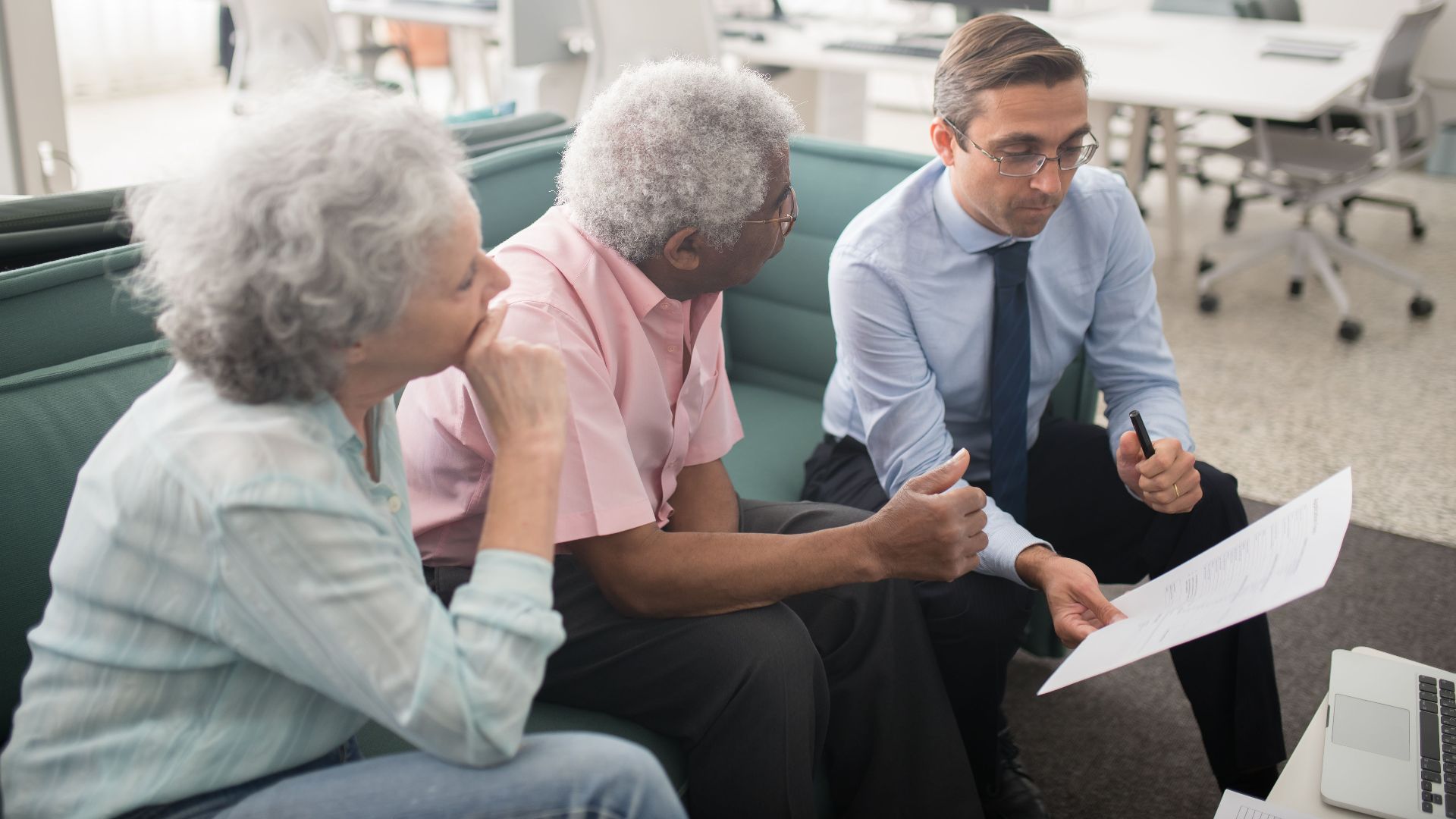 Business professional consults elderly clients in an office setting. Collaborative discussion, paperwork visible.