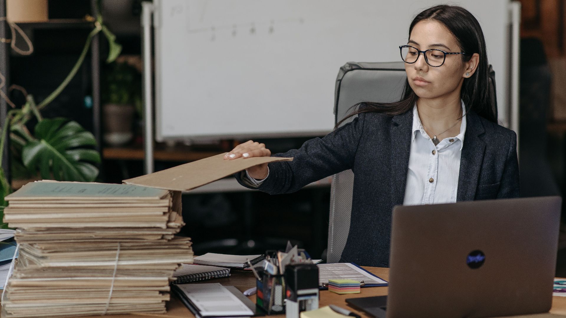 A businesswoman in a modern office organizing a stack of files beside a laptop.