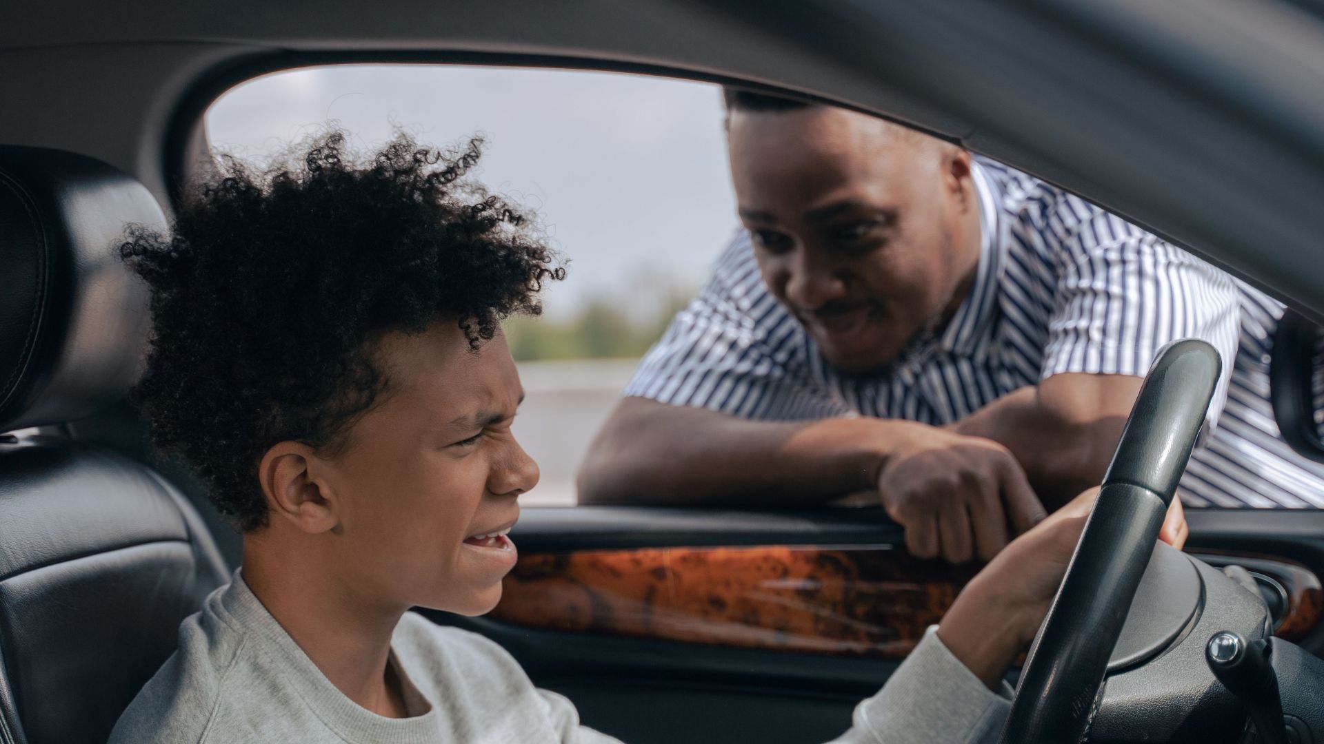Father instructs son in car driving lesson inside a vehicle on a sunny day.