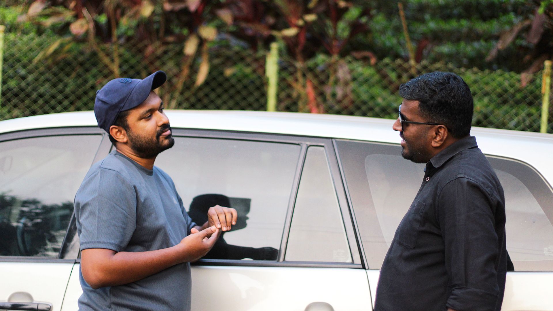 Two men having a casual discussion beside a car in an outdoor setting.