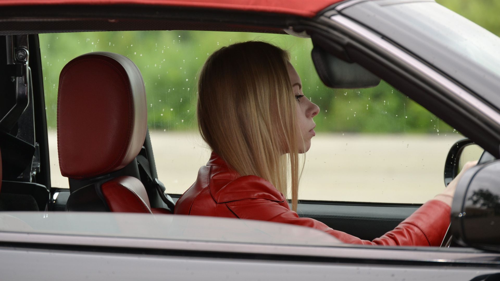 A young woman in a red convertible driving on a rainy day, wearing a red jacket.
