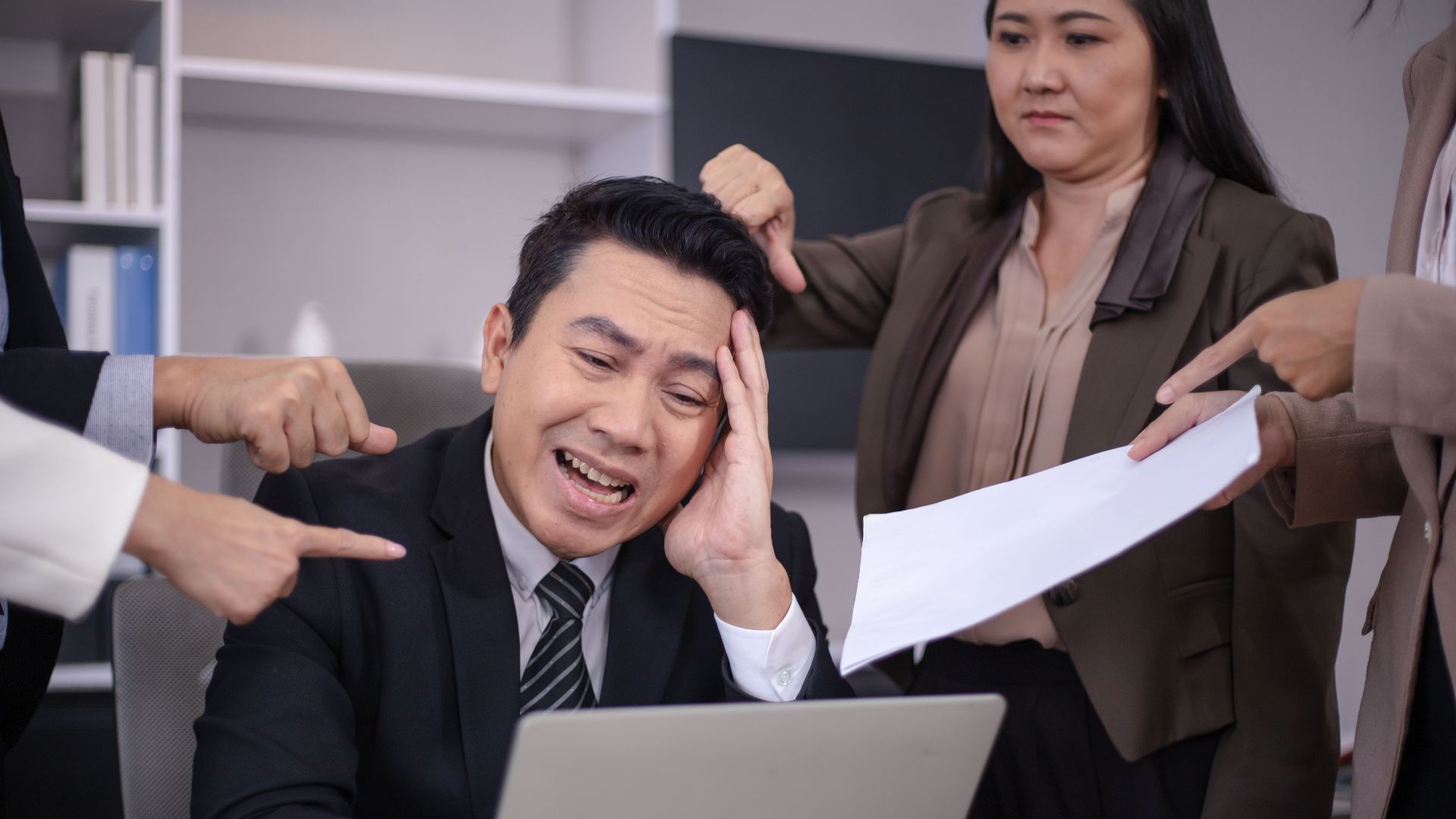 An Asian businessman looks stressed as colleagues point fingers and give him paperwork in a modern office.