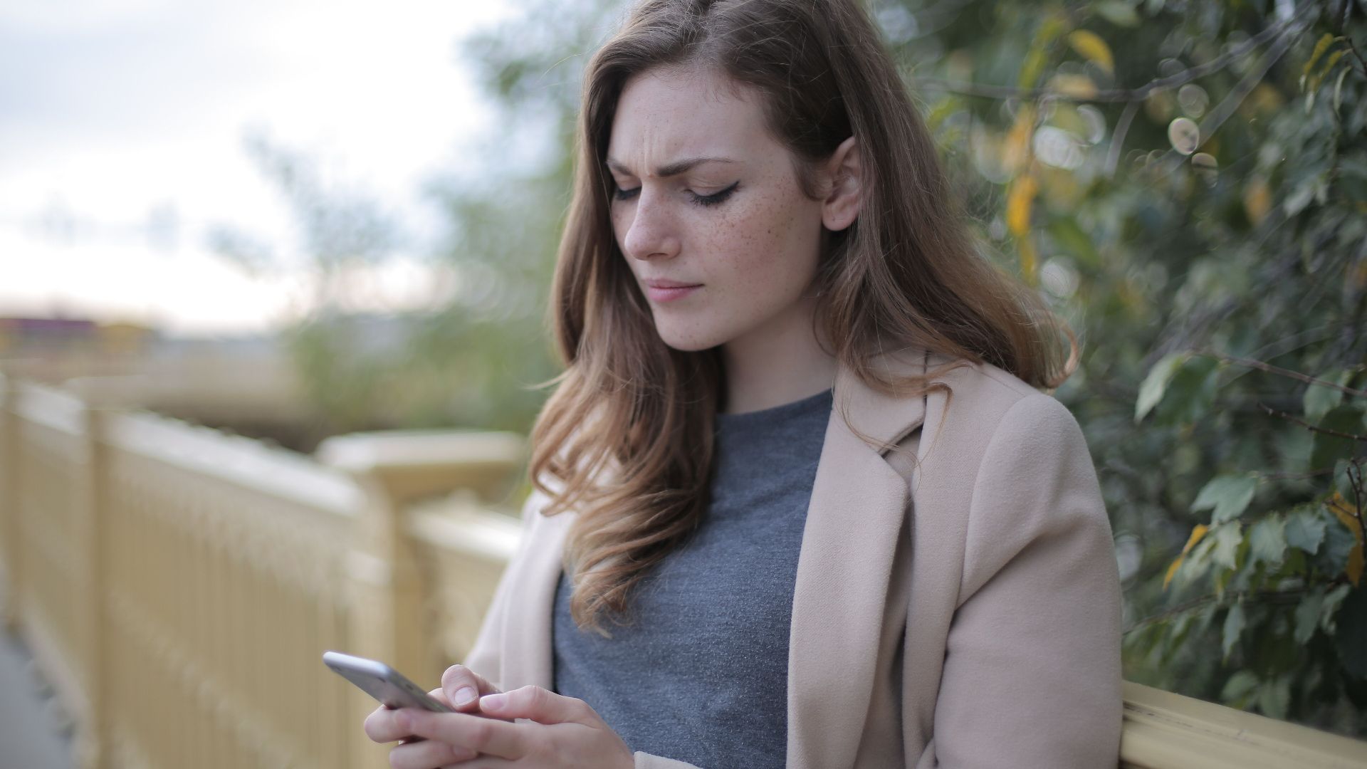A woman stands outdoors in a casual outfit, using her smartphone with a pensive look.