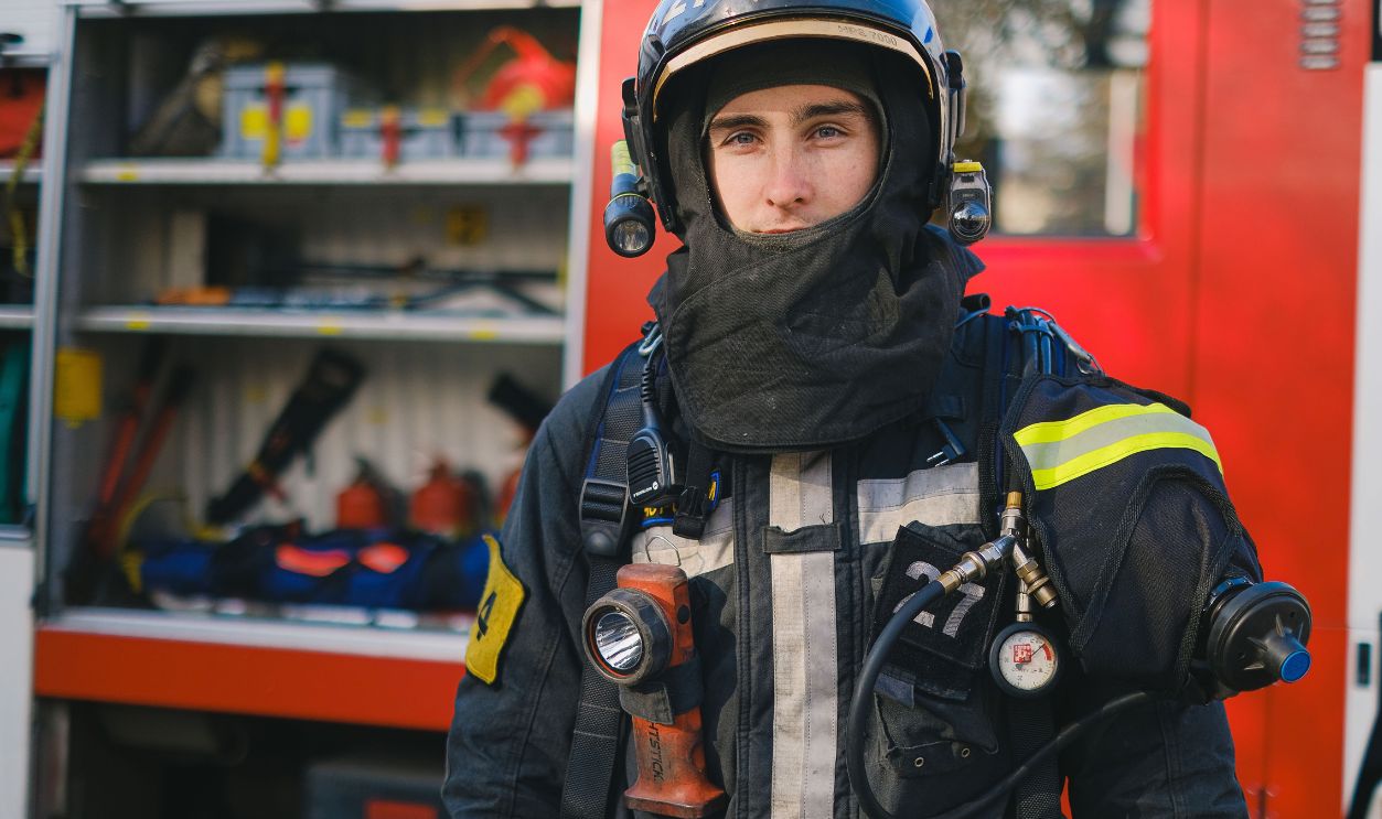 A firefighter stands in front of a fire truck wearing full protective gear and helmet