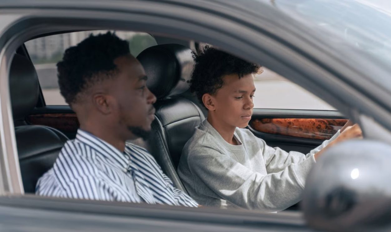 A father sits beside his son who is learning to drive, inside a car