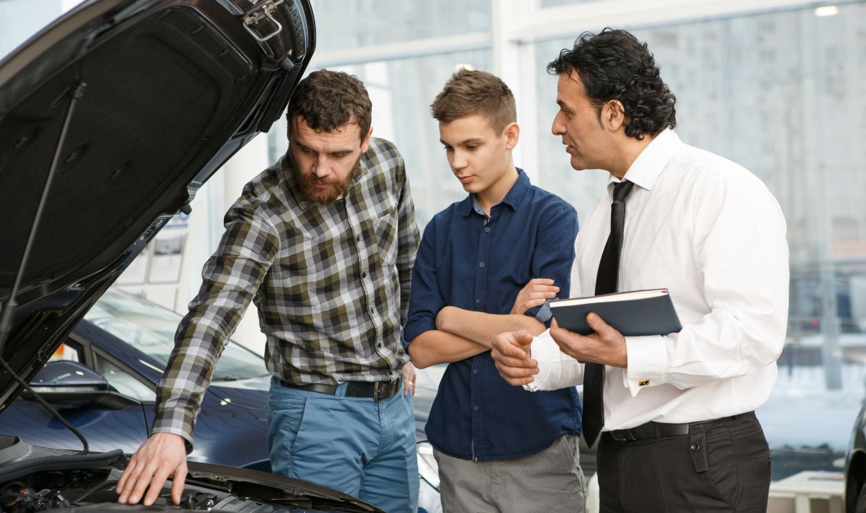 Handsome mature man and his teenage son choosing a new car at the dealership talking to a professional salesman checking out engine of a car with a n open hood automotive cars sales vehicle transport