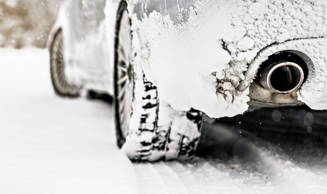 Silver car covered with ice parked on snow covered road, detail view to exhaust from behind, blurred trees background empty space for text left side