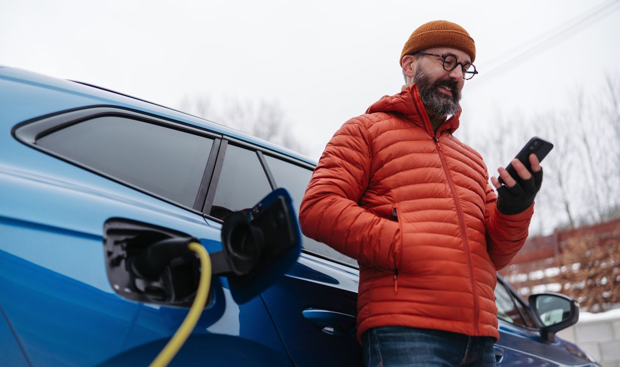Man charging electric car during cold snowy day, using electric vehicle charging app, checking battery life, energy consumption on smart phone. Charging and driving electric vehicles during winter