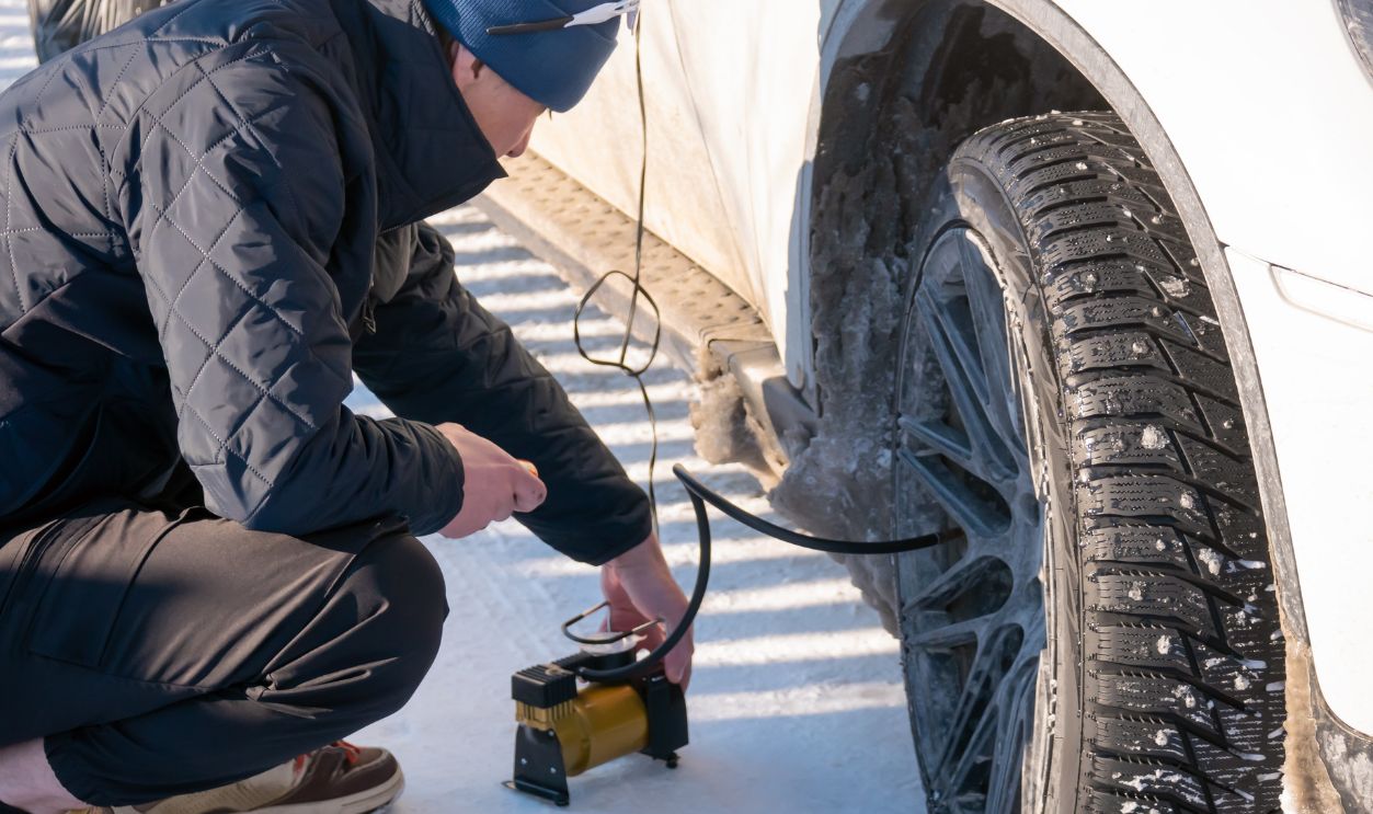 Man inflating car tire with portable electric pump on snowy road. Driver checking tire pressure in wintertime. Vehicle maintenance and emergency roadside assistance during cold weather.