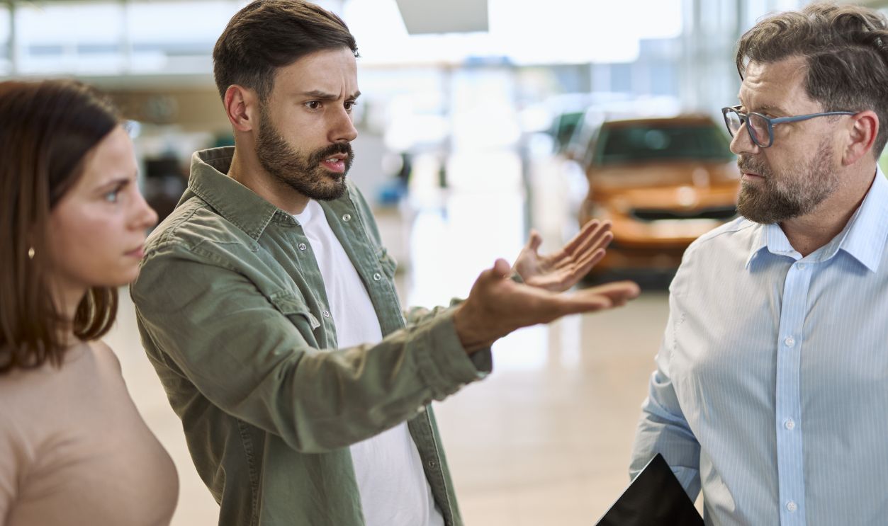Displeased man and his wife arguing with car salesperson in a car showroom.