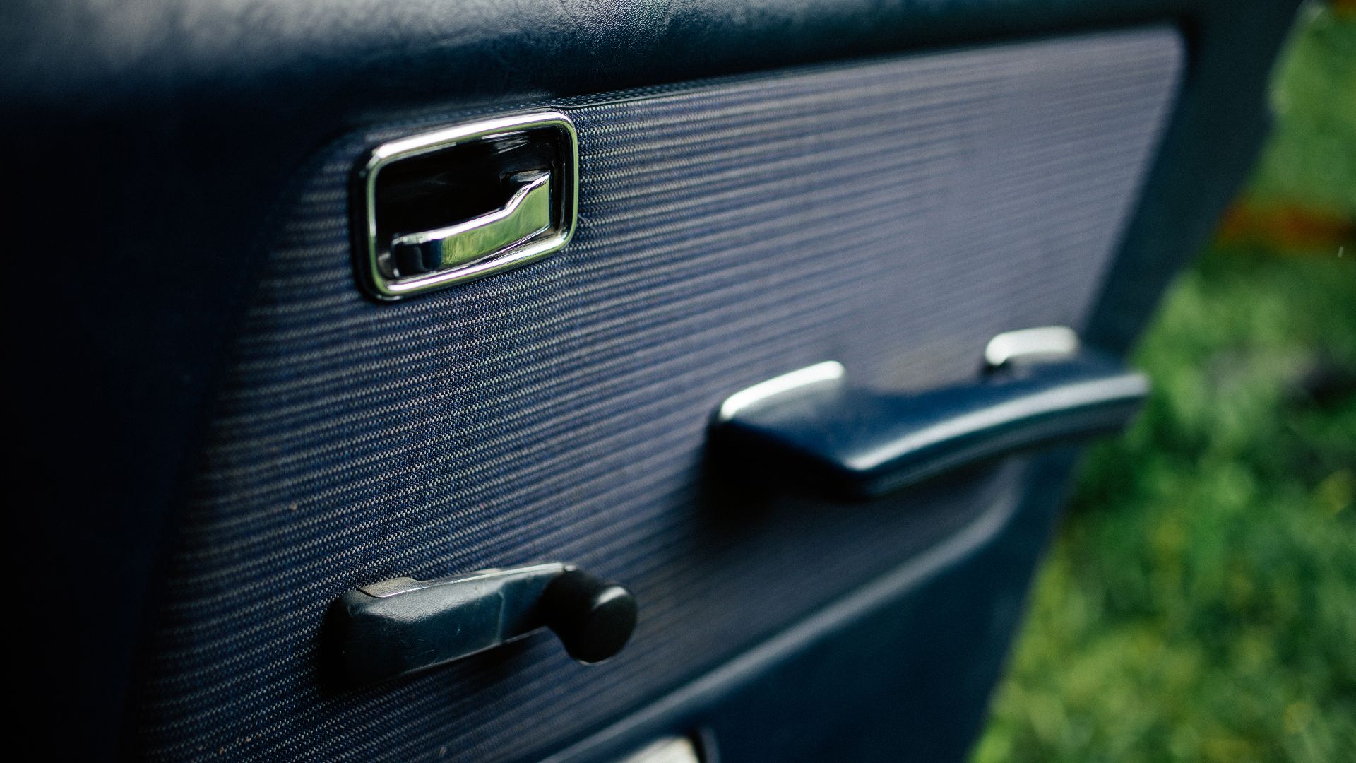 A classic car shows its door handle and window adjustment crank. Sunlight highlights their design. Lush green grass is visible in the background.