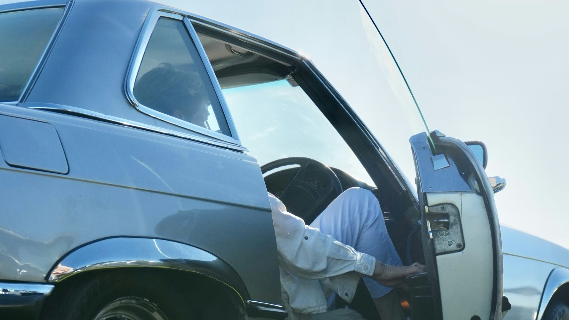 A person in a white shirt and trousers exiting a silver vintage car