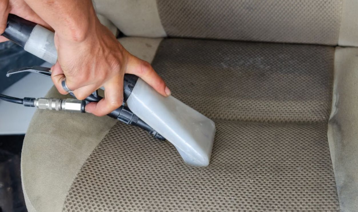 Hands using a vacuum nozzle to deeply clean a fabric car seat, removing dirt and stains