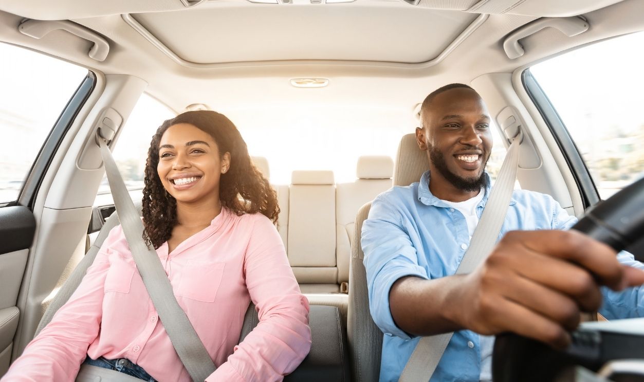 Front view portrait of smiling handsome African American man driving modern luxury car, happy beautiful young woman sitting on the front passenger seat, looking on the road, sun flare