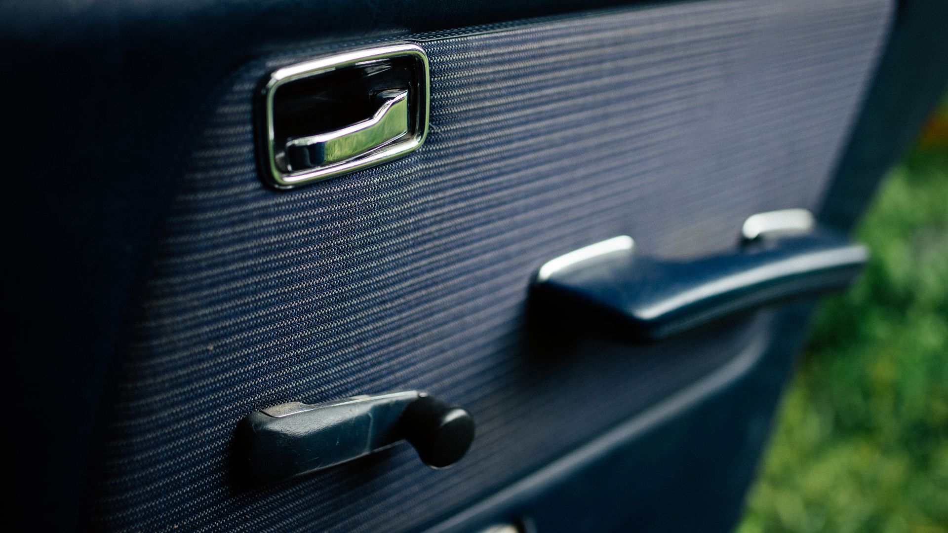 A classic car shows its door handle and window adjustment crank. Sunlight highlights their design. Lush green grass is visible in the background.