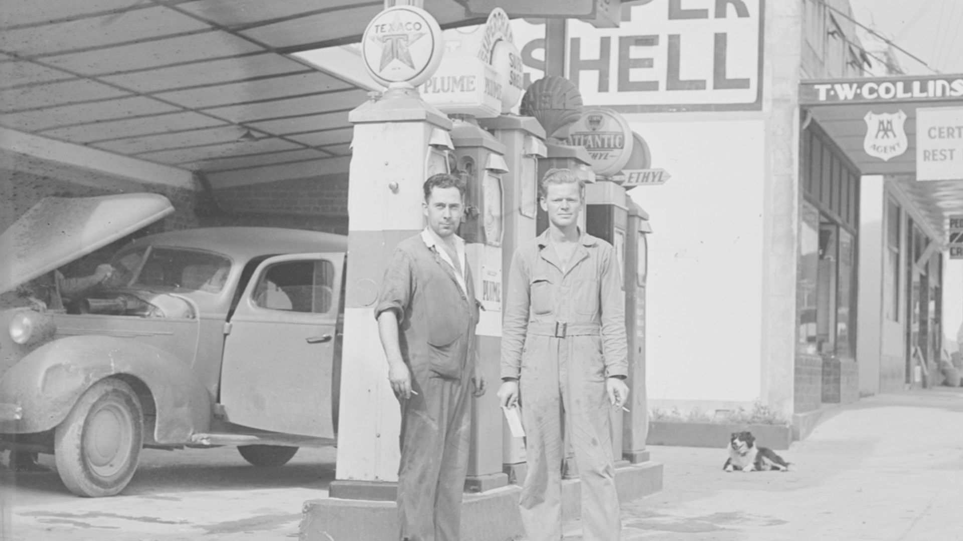 T. W. Collins Warkworth Service Station with portrait of two service station men- attendants. There are multiple signs on the petrol pumps,