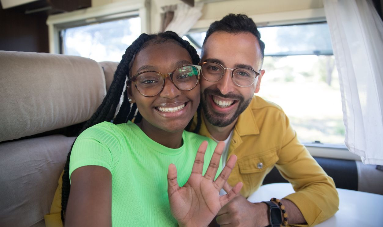Happy couple smiling and waving inside an RV, enjoying a road trip adventure.
