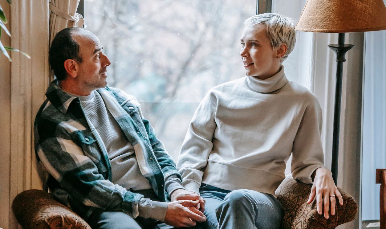 A couple sits closely on a couch by the window, enjoying a cozy and relaxed moment indoors.