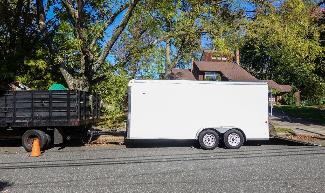 A landscaping truck with a long white enclosed trailer trailer seen on a shady residential asphalt street