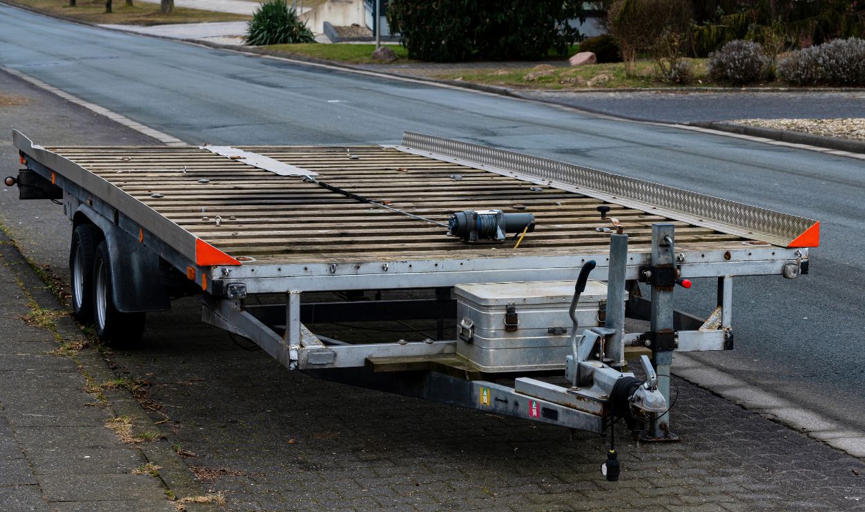 A parked transport trailer rests on the roadside, featuring a wooden deck and metal frame. The surroundings show a calm street with minimal traffic and greenery nearby.