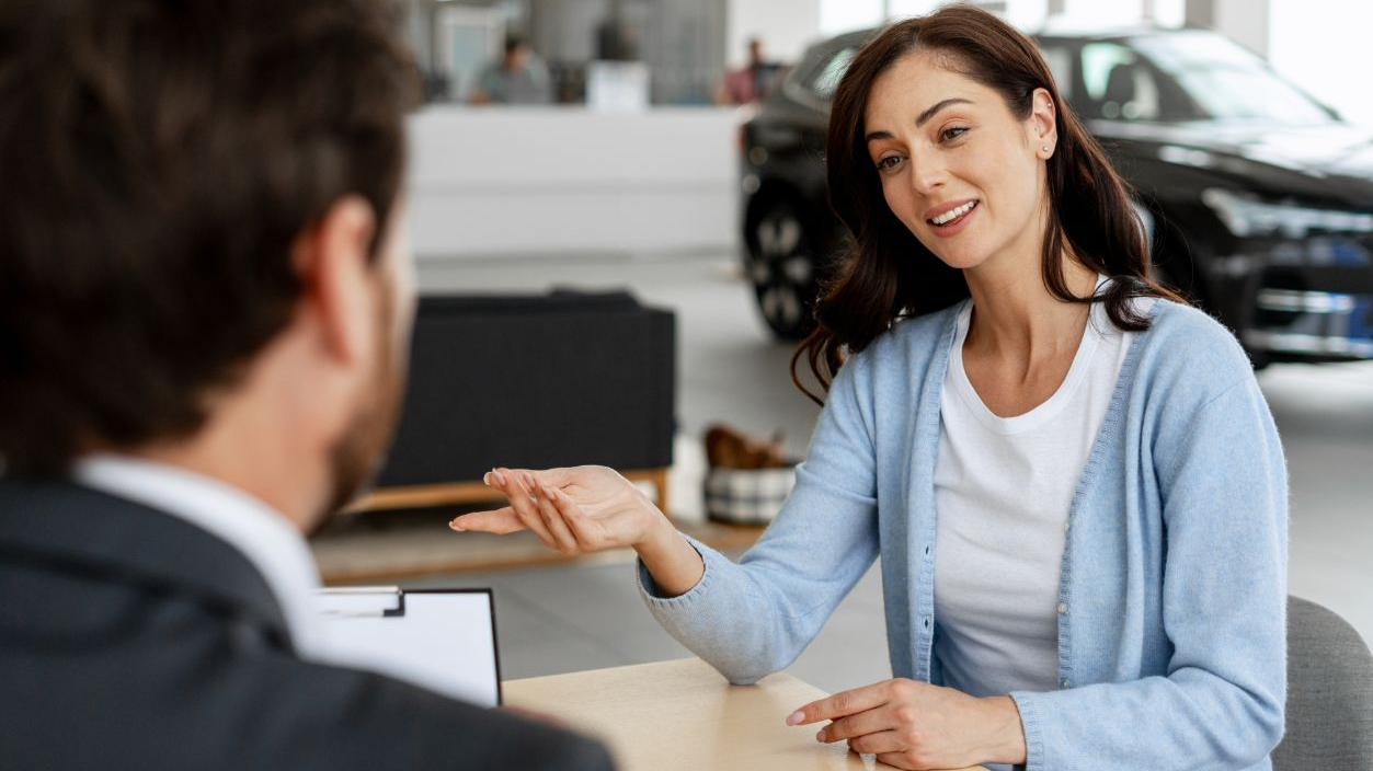 Shutterstock-2657401983, Customer discussing her needs with a car dealer while seated at a desk in a bustling dealership, navigating the buying process together