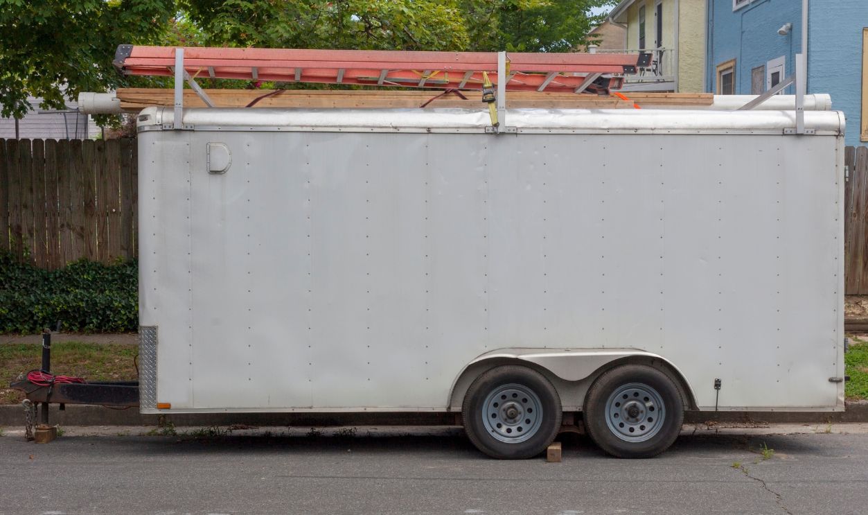 Side view of older white utility work trailer parked in residential neighborhood with ladders on top.