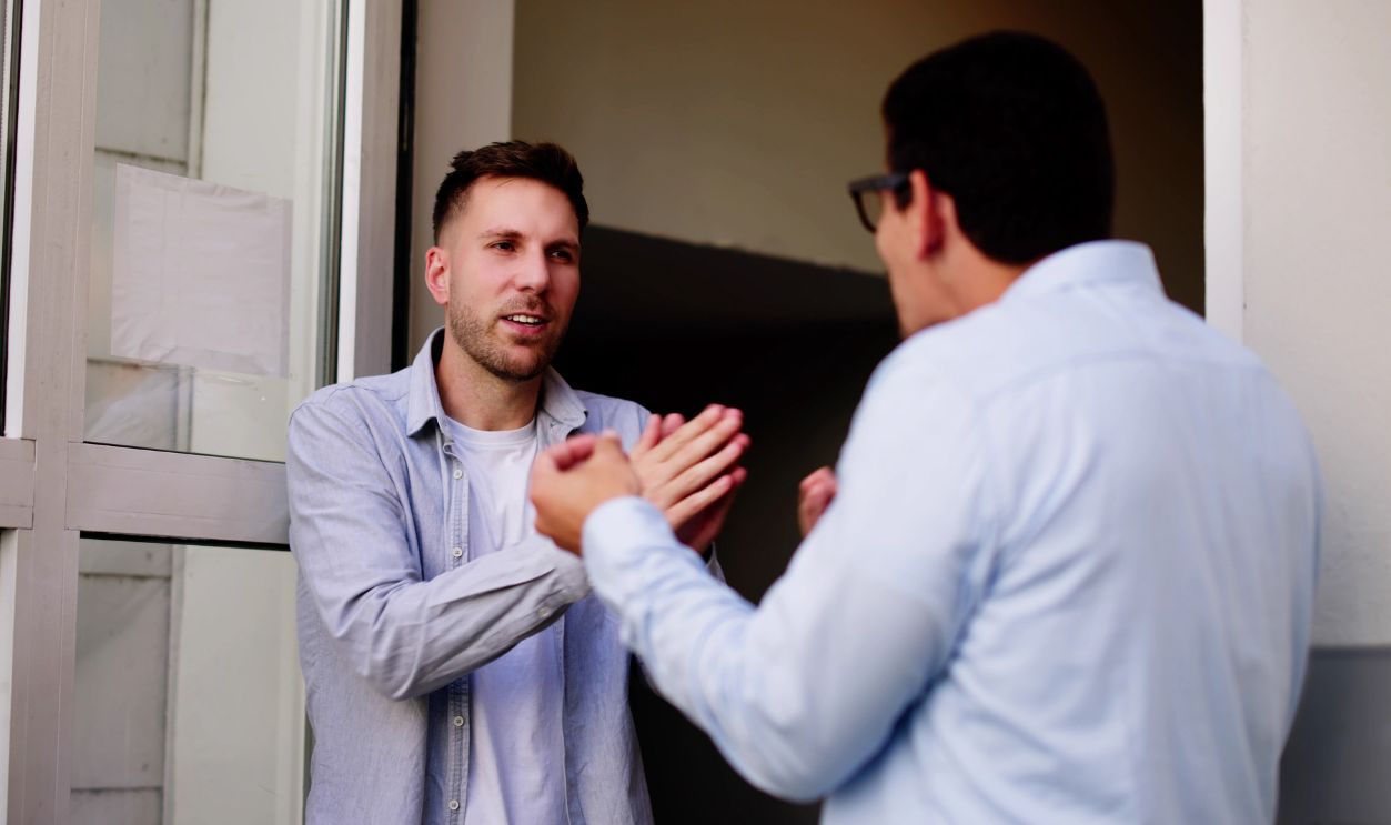 Two young men, one Hispanic, one Caucasian, arguing loudly at neighbor's door entrance, pointing fingers and shrugging shoulders.