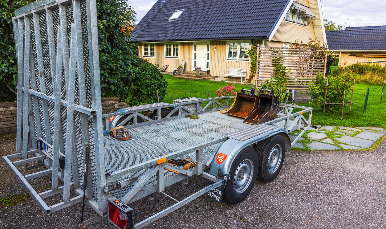 Metal trailer with excavator buckets parked on driveway in front of residential house with garden and green lawn. Sweden. Stockholm. 09.10.2025.