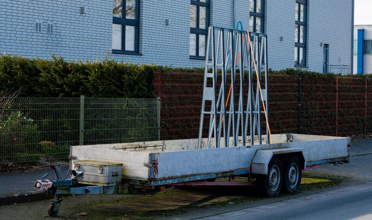 A car trailer stands stationary near a building. The trailer is equipped with a simple framework, situated on a street with greenery along the fence in an urban area during daylight.