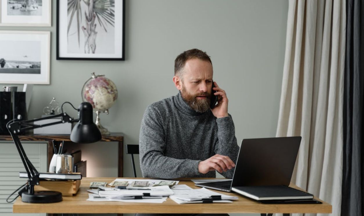 A Bearded Man Using a Laptop Near Cash Money on Wooden Table
