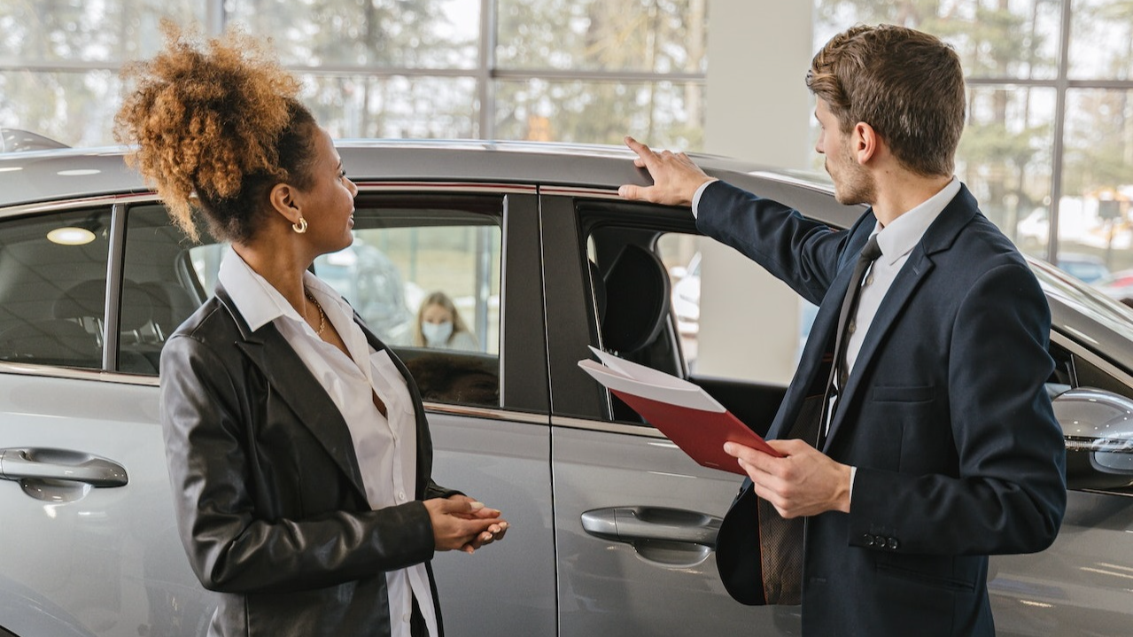 A woman is talking with a man wearing black suit in car dealership.