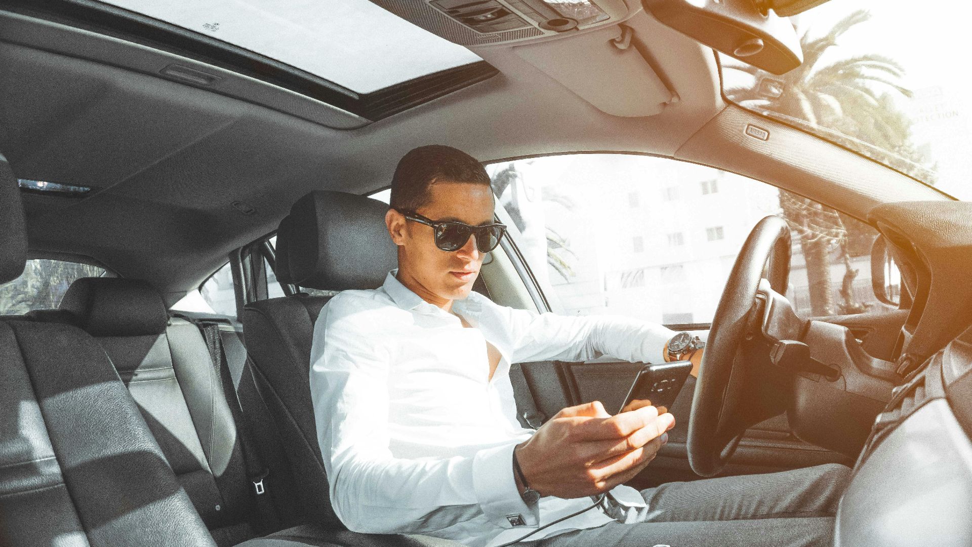 A man wearing sunglasses texts on his phone while driving a car in Morocco.
