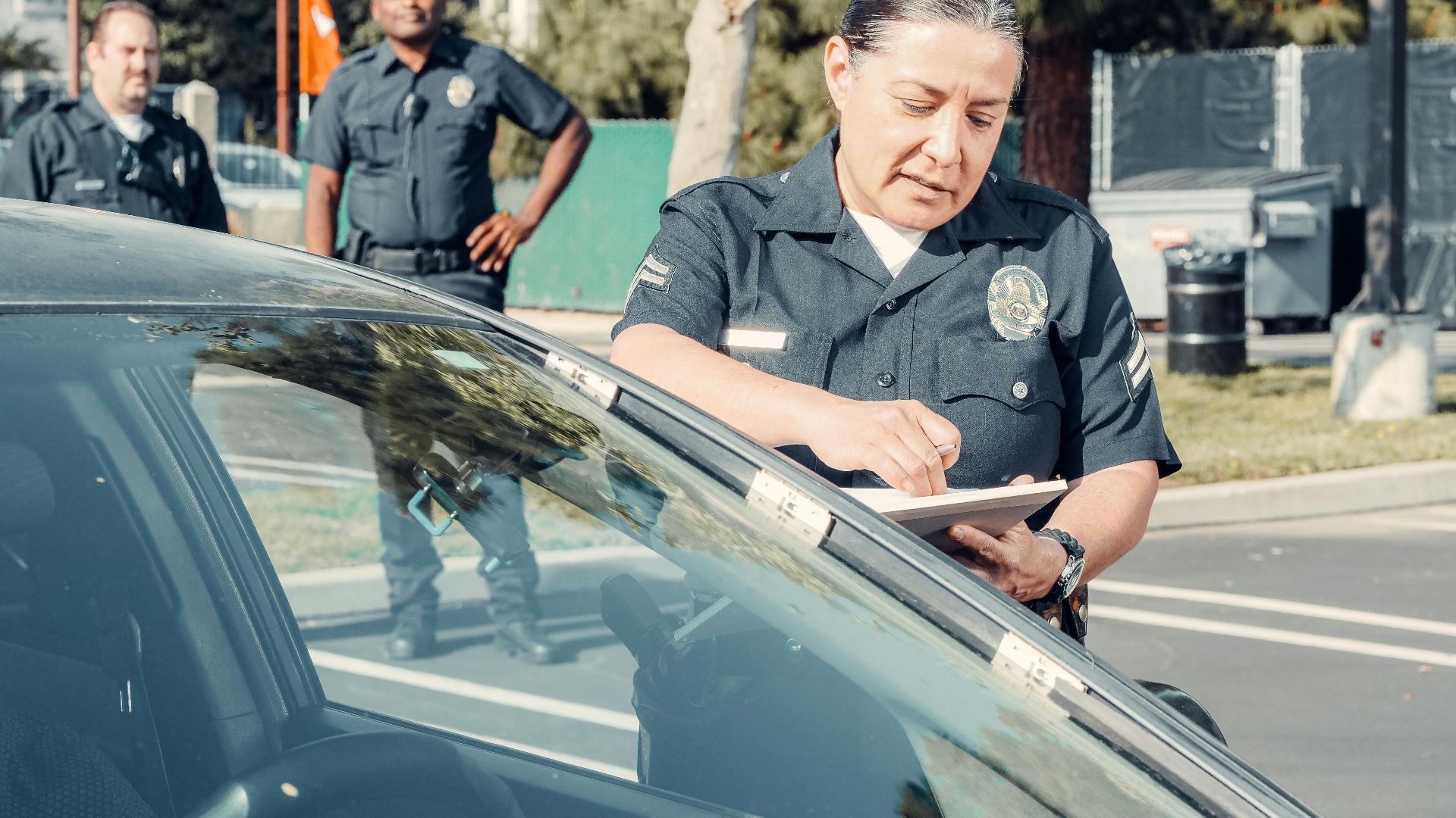 Police officers handling a traffic violation in a parking lot, focus on policewoman writing a ticket.