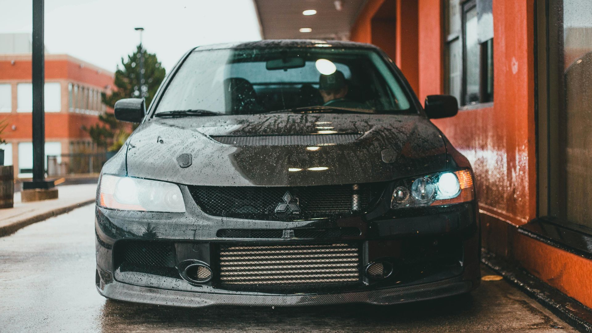 A black car parked on a wet street, reflecting rain near an orange building.