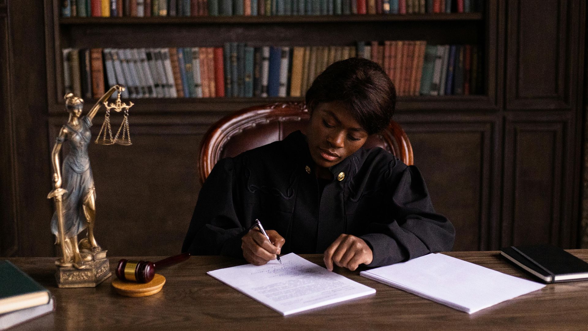 A judge in robes writing on a document at a desk in an office library with law books.
