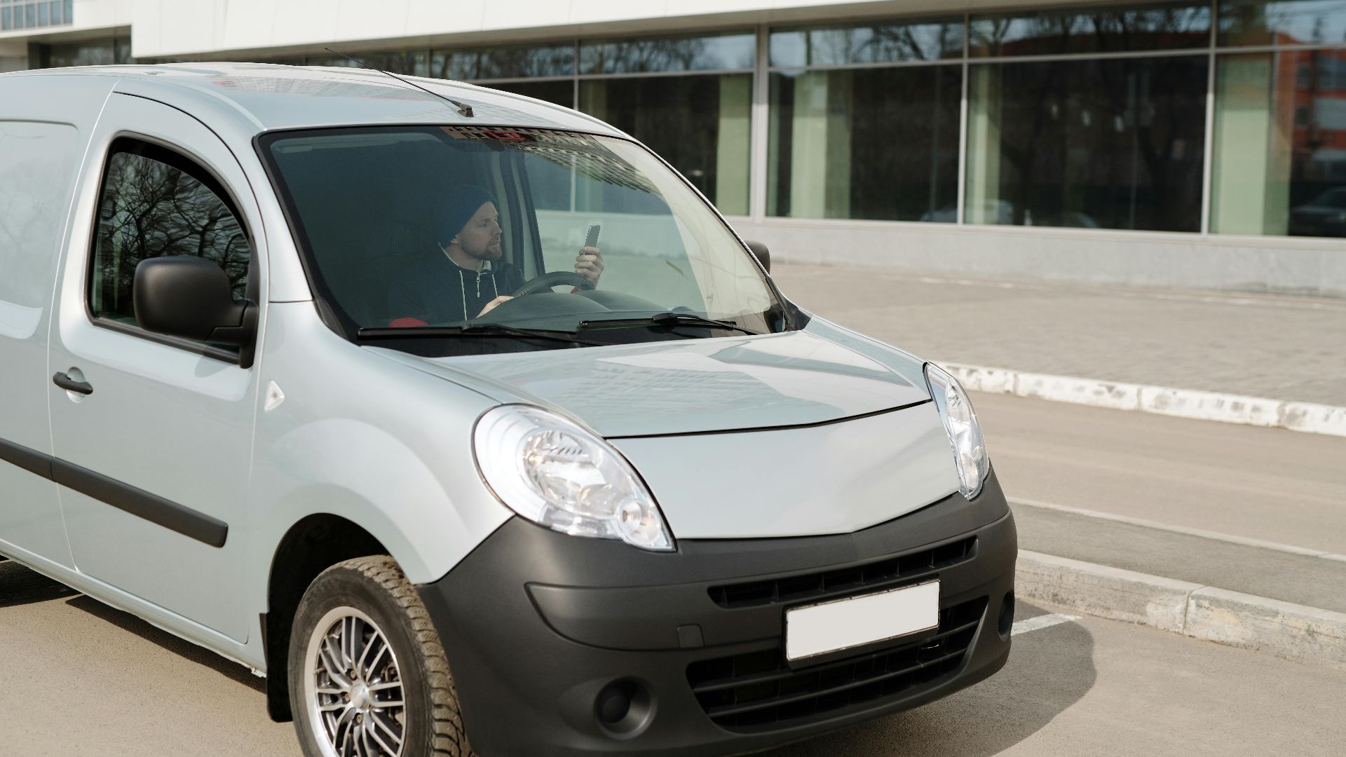 A delivery driver in a van using a smartphone while parked outside a modern building.