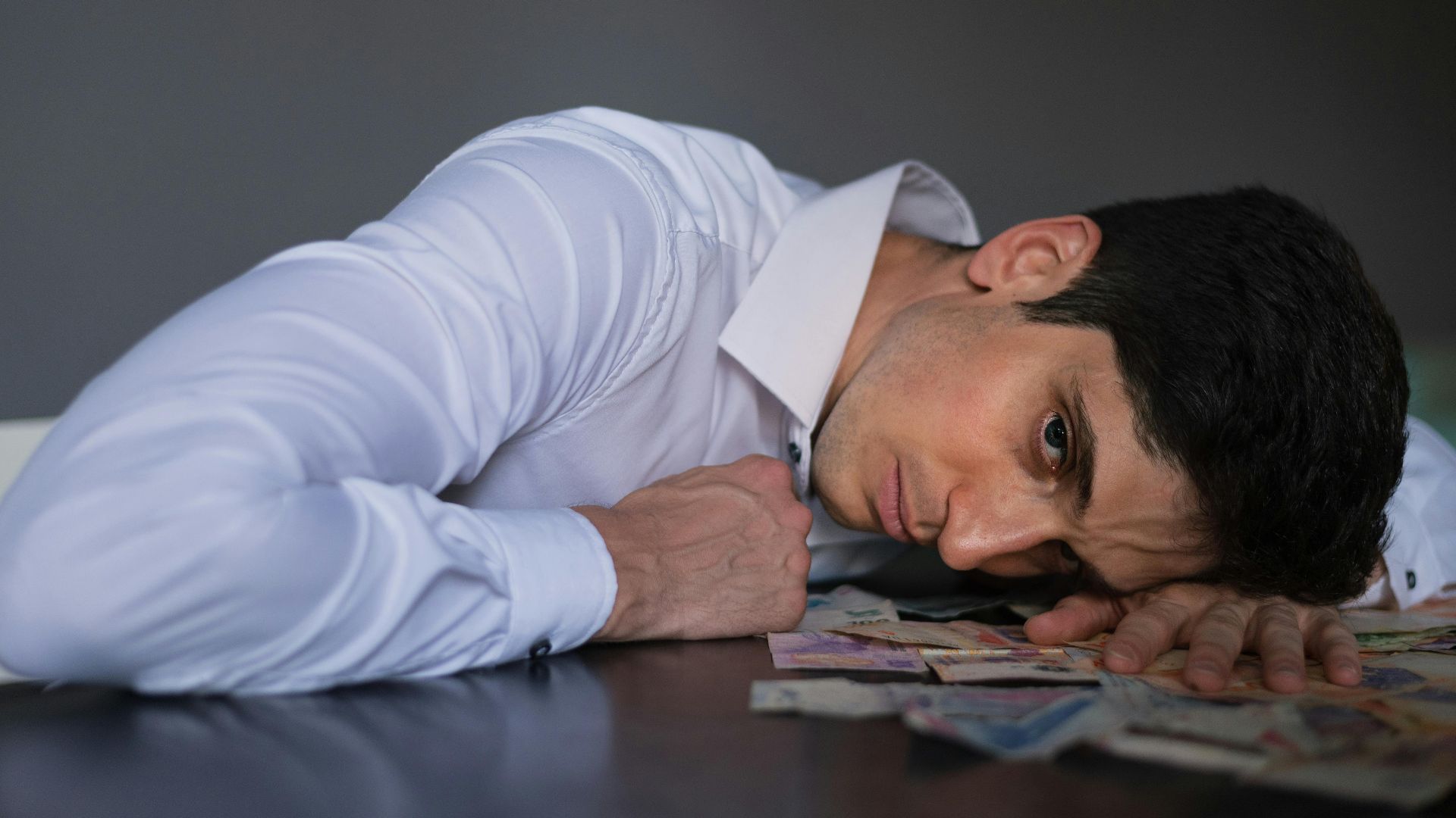 Adult man in shirt lying on table, surrounded by currency, looking stressed indoors.