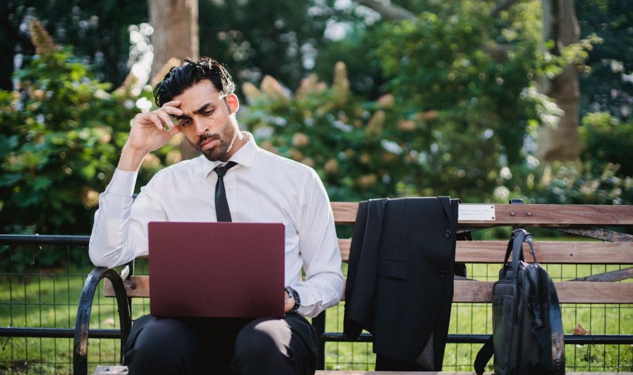 A Man in a White Long Sleeves and Black Pants Using His Laptop While Sitting on a Brown Wooden Bench