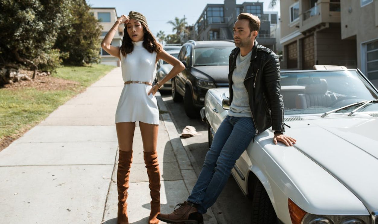 Woman in White Dress and Man in Black Leather Jacket on Sidewalk Near Cars