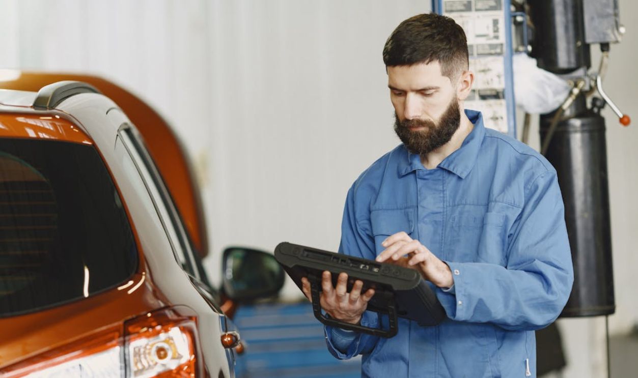 Man in Blue Coveralls Standing Beside an Orange Car Using an Automotive Diagnostic Tool