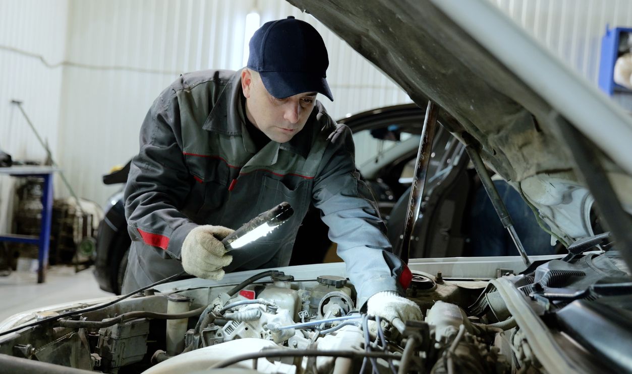 Professional mechanic inspecting vehicle engine, wearing protective gear and using flashlight while performing detailed diagnostic work inside modern automotive repair facility