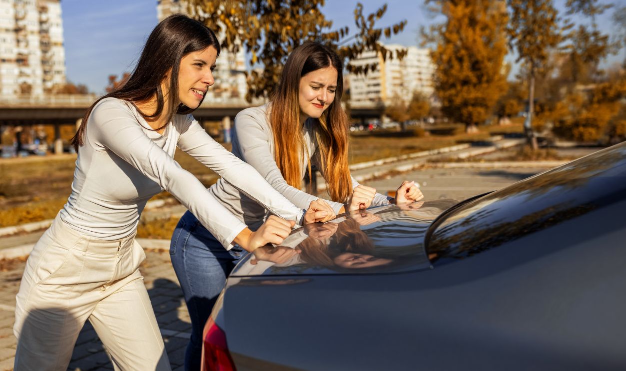 Two young women pushing a car that won’t start, working together on the roadside, daylight, casual everyday moment.