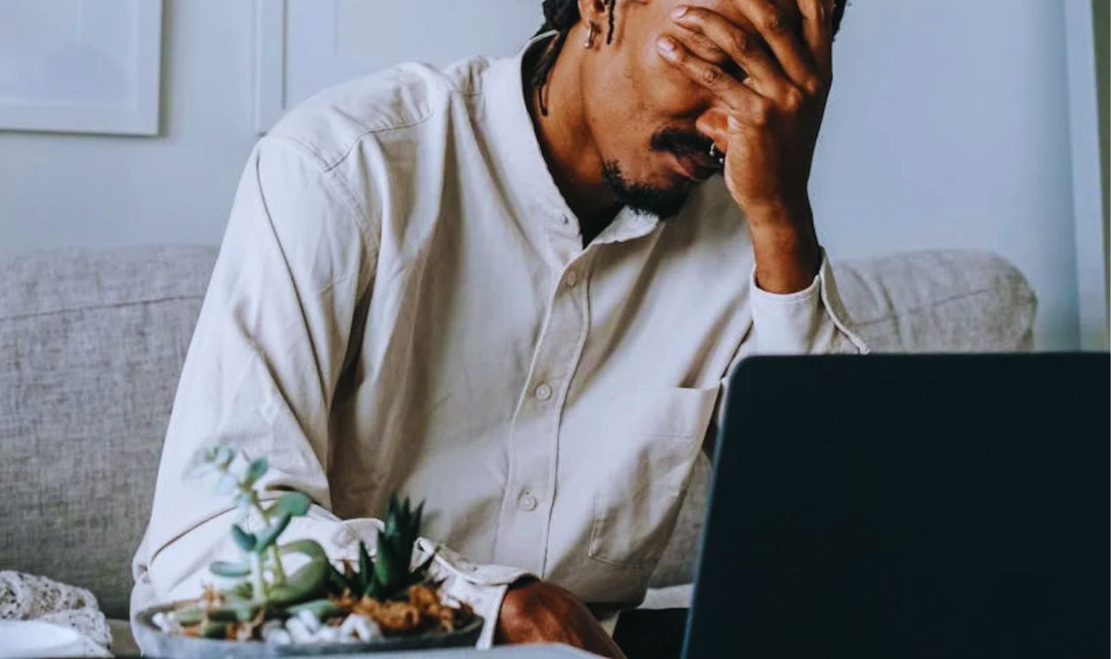 Upset young black guy covering face with hand while working remotely on netbook