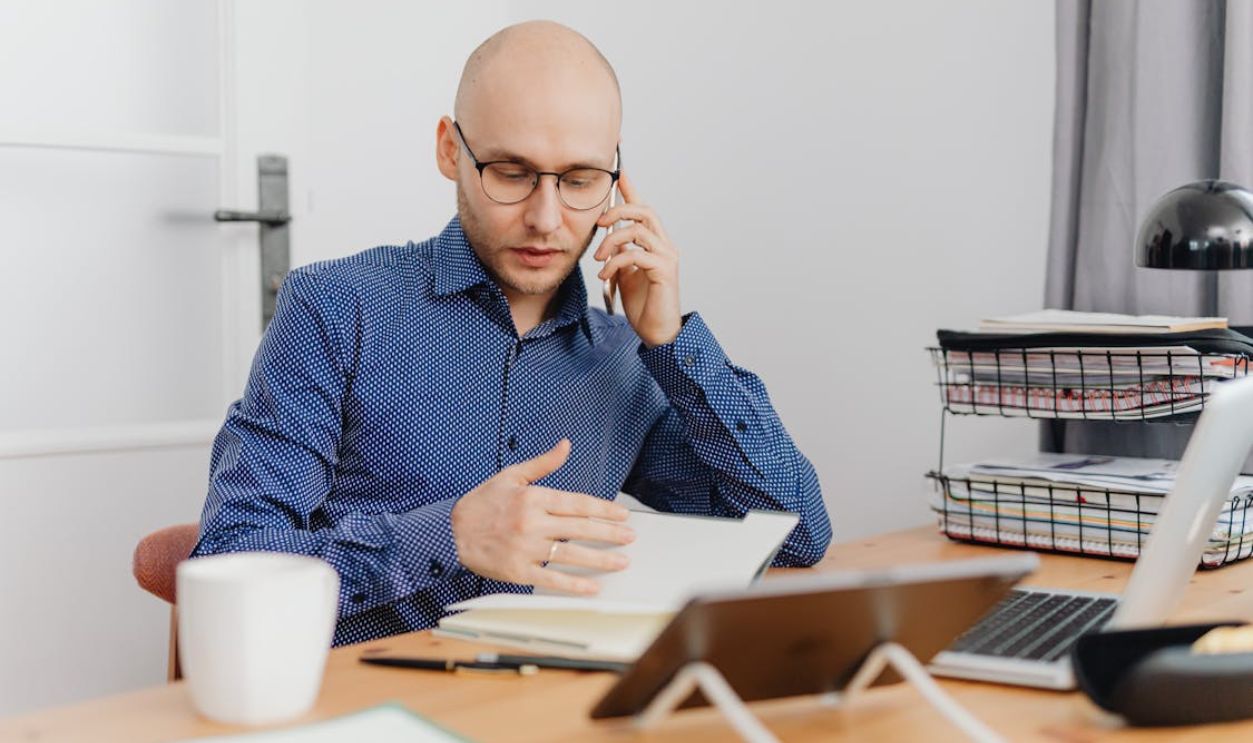 Man in Blue and White Polka Dots Dress Shirt Using Cellphone While Looking At His Notebook